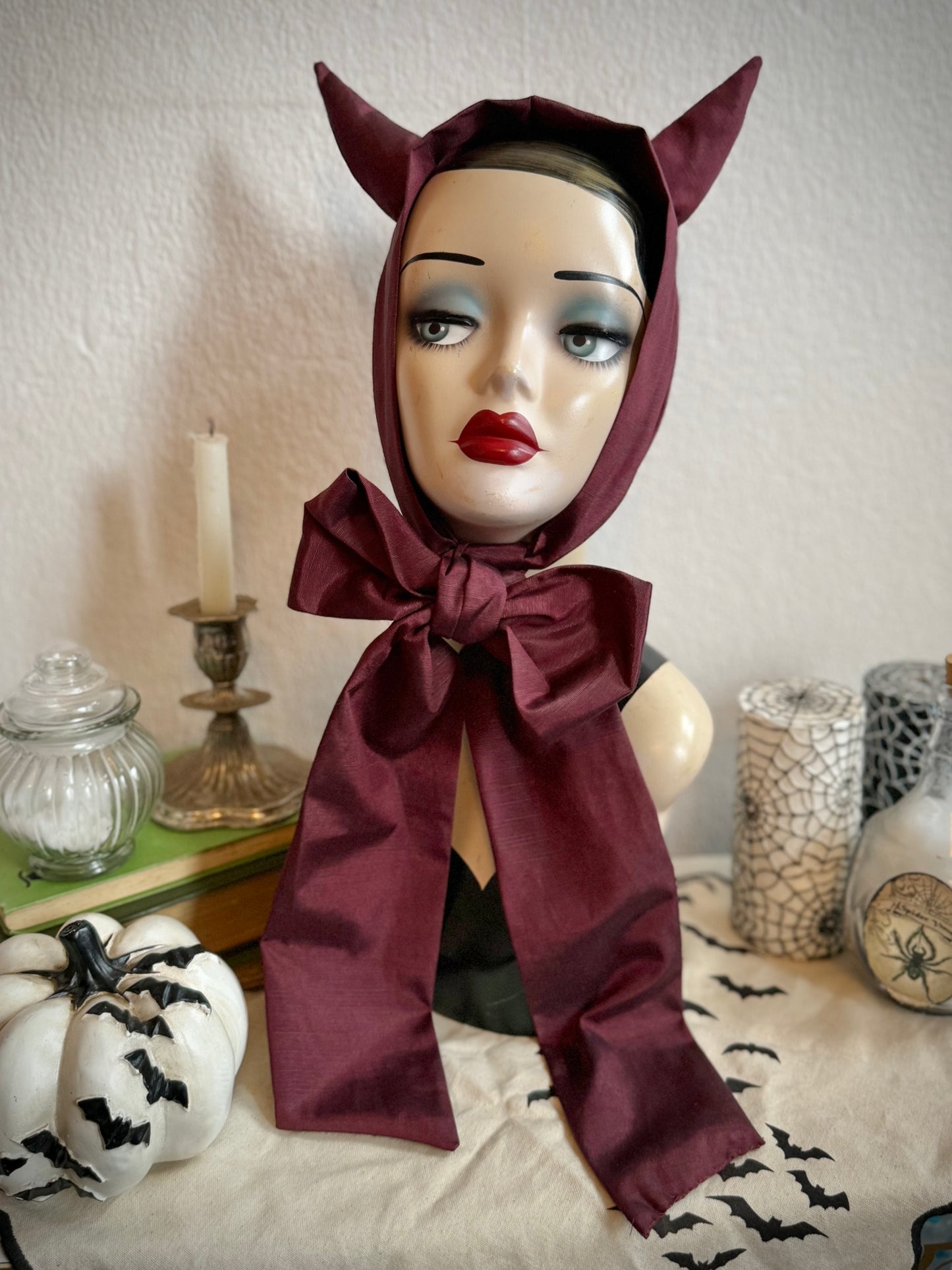 Mannequin head wearing a red devil horns headpiece with a large bow, surrounded by Halloween decorations.