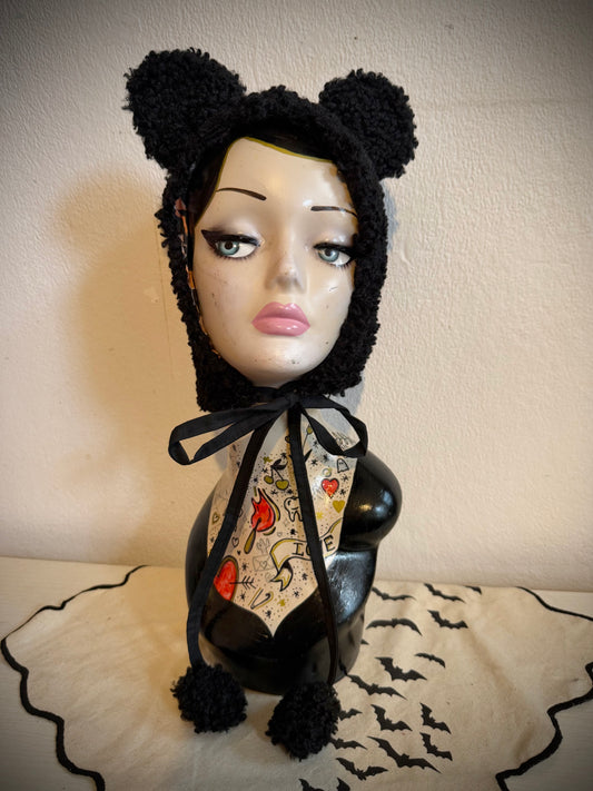 Black fuzzy bear-ear bonnet with a black ribbon bow and pom-poms, displayed on a mannequin bust against a beige wall.